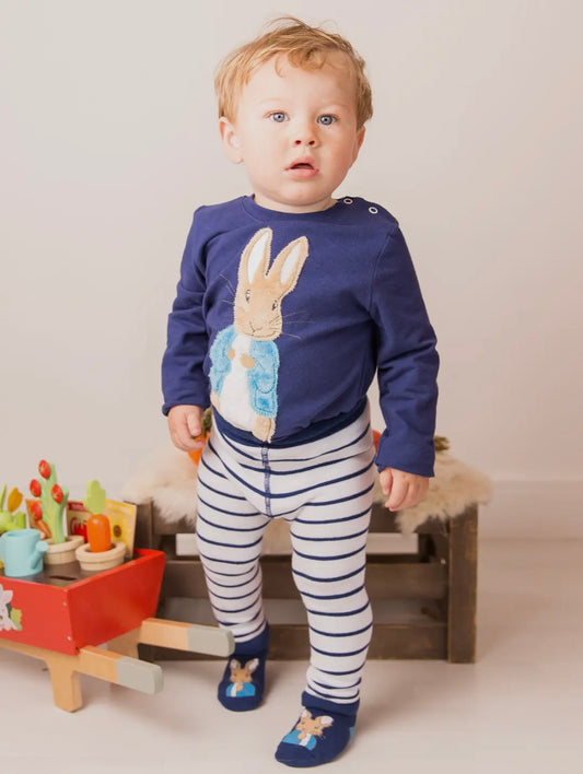 Child wearing a navy blue Peter Rabbit motif with a bunny design, striped pants, and matching shoes, standing in a room with toys.