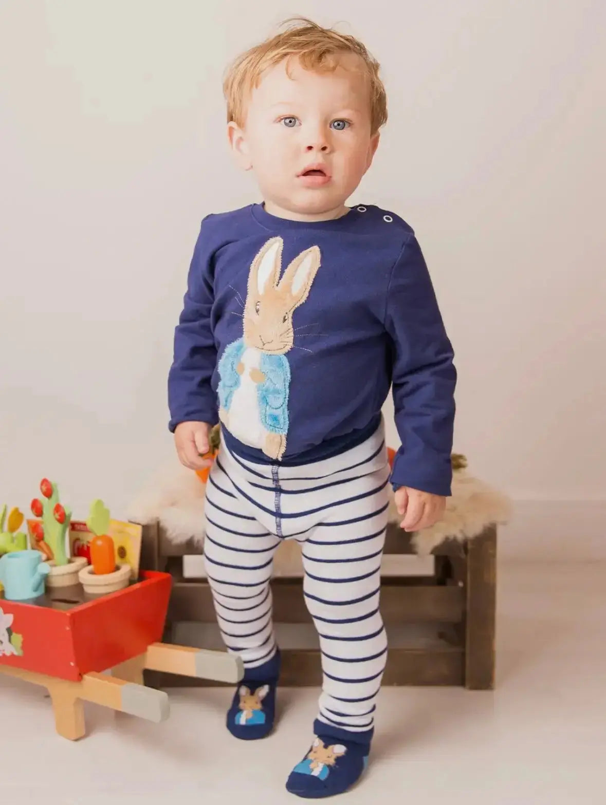 Child wearing a navy blue Peter Rabbit motif with a bunny design, striped pants, and matching shoes, standing in a room with toys.